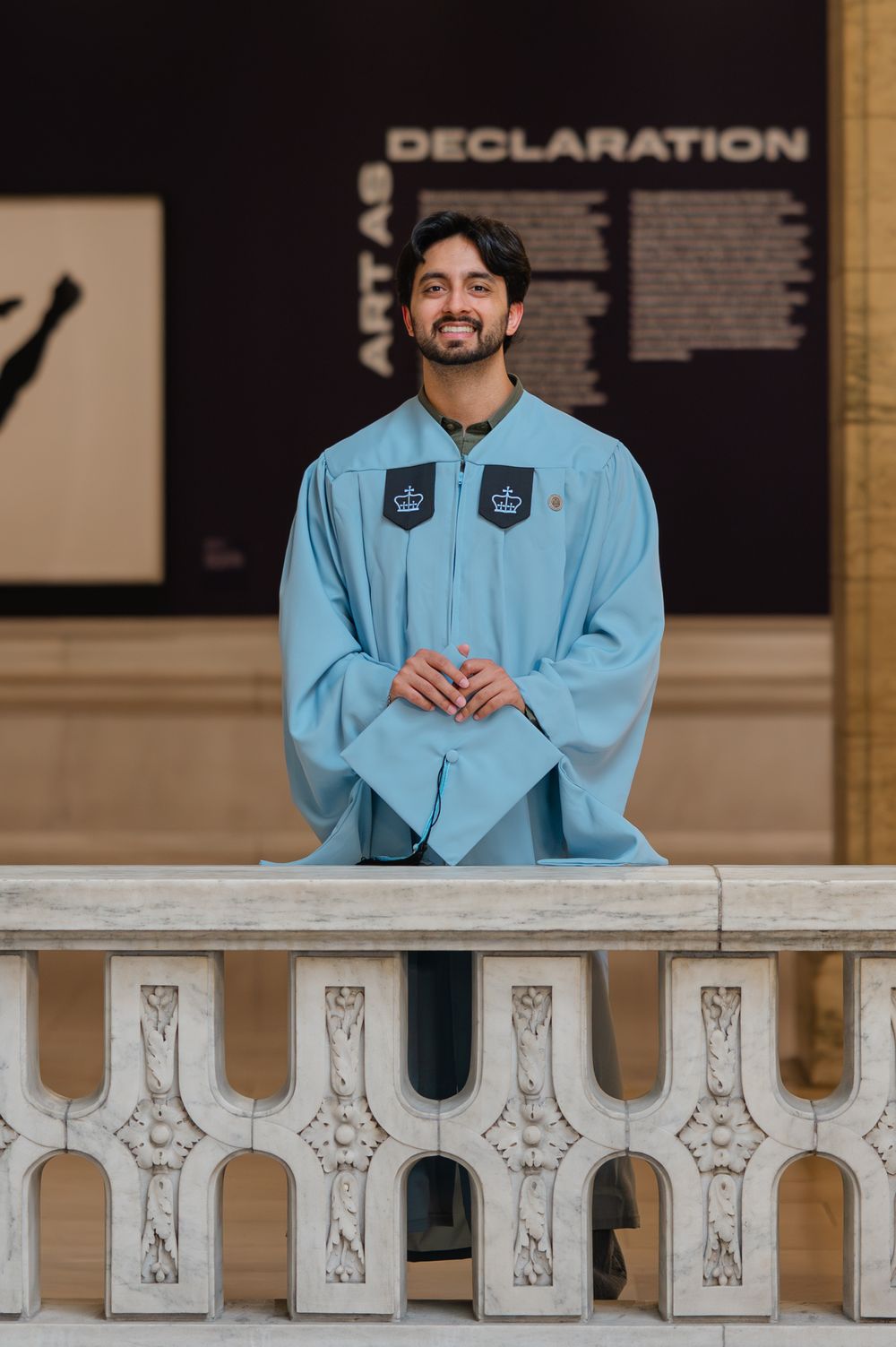 Smiling graduate in light blue graduation gown stands at ornate balcony inside museum with Declaration exhibit backdrop.