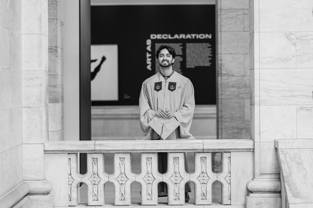 Graduate in cap and gown stands on balcony before Declaration sign in black and white.