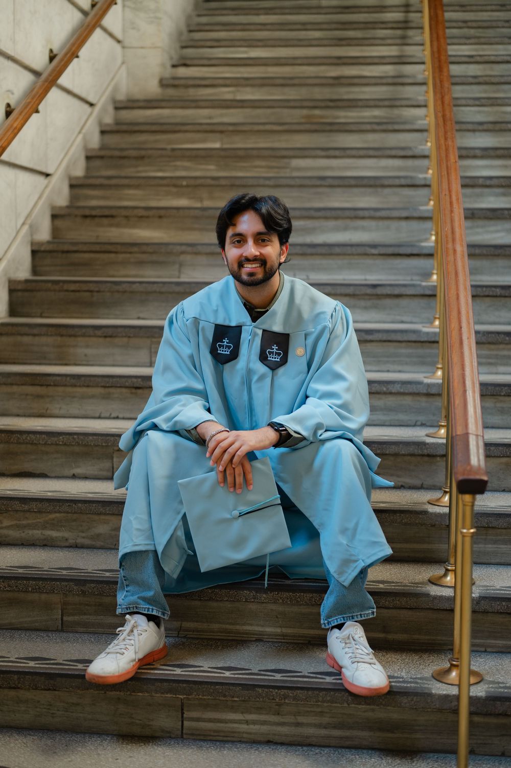 Young man in light blue graduation gown sitting on stone steps, smiling, holding cap, wearing white sneakers.