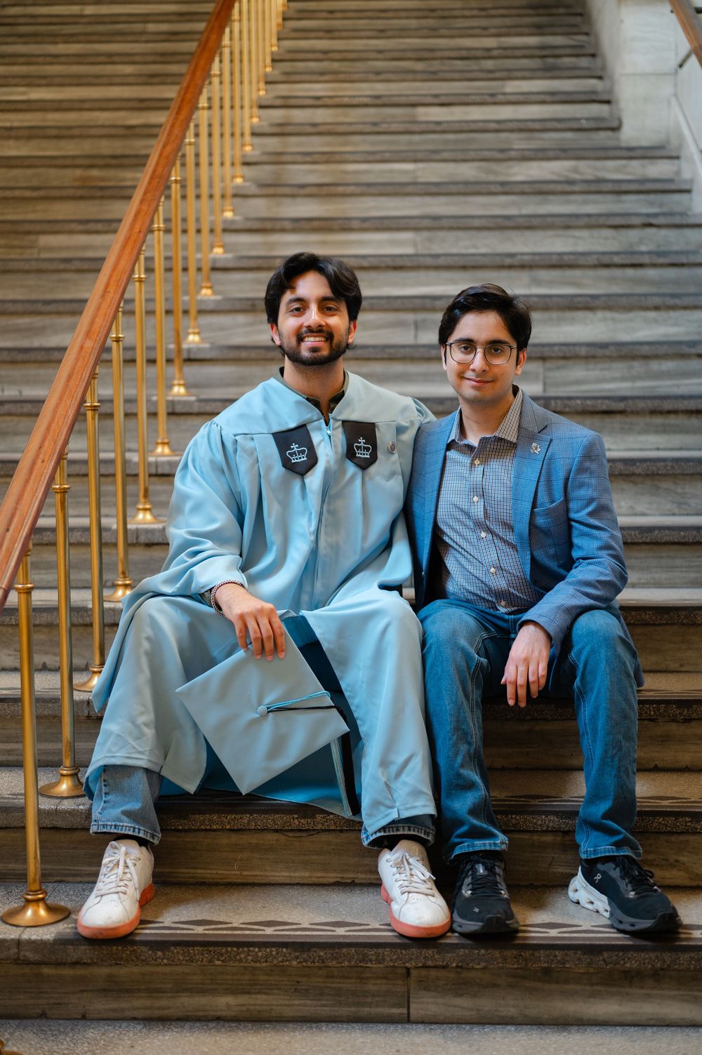 Two young men sit on marble stairs, one wearing a light blue graduation robe and cap.