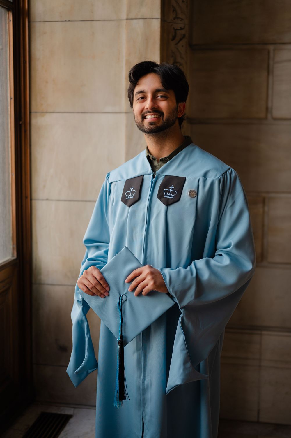 Smiling graduate in light blue cap and gown holding mortarboard at graduation ceremony.