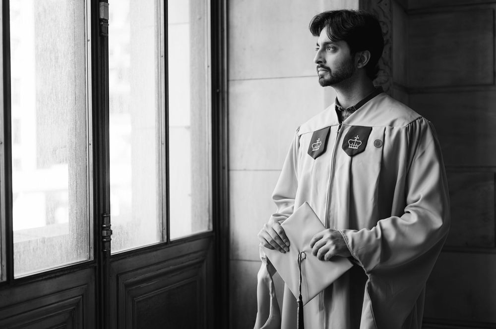 Man in graduation gown holding documents stands by window in black and white photo.