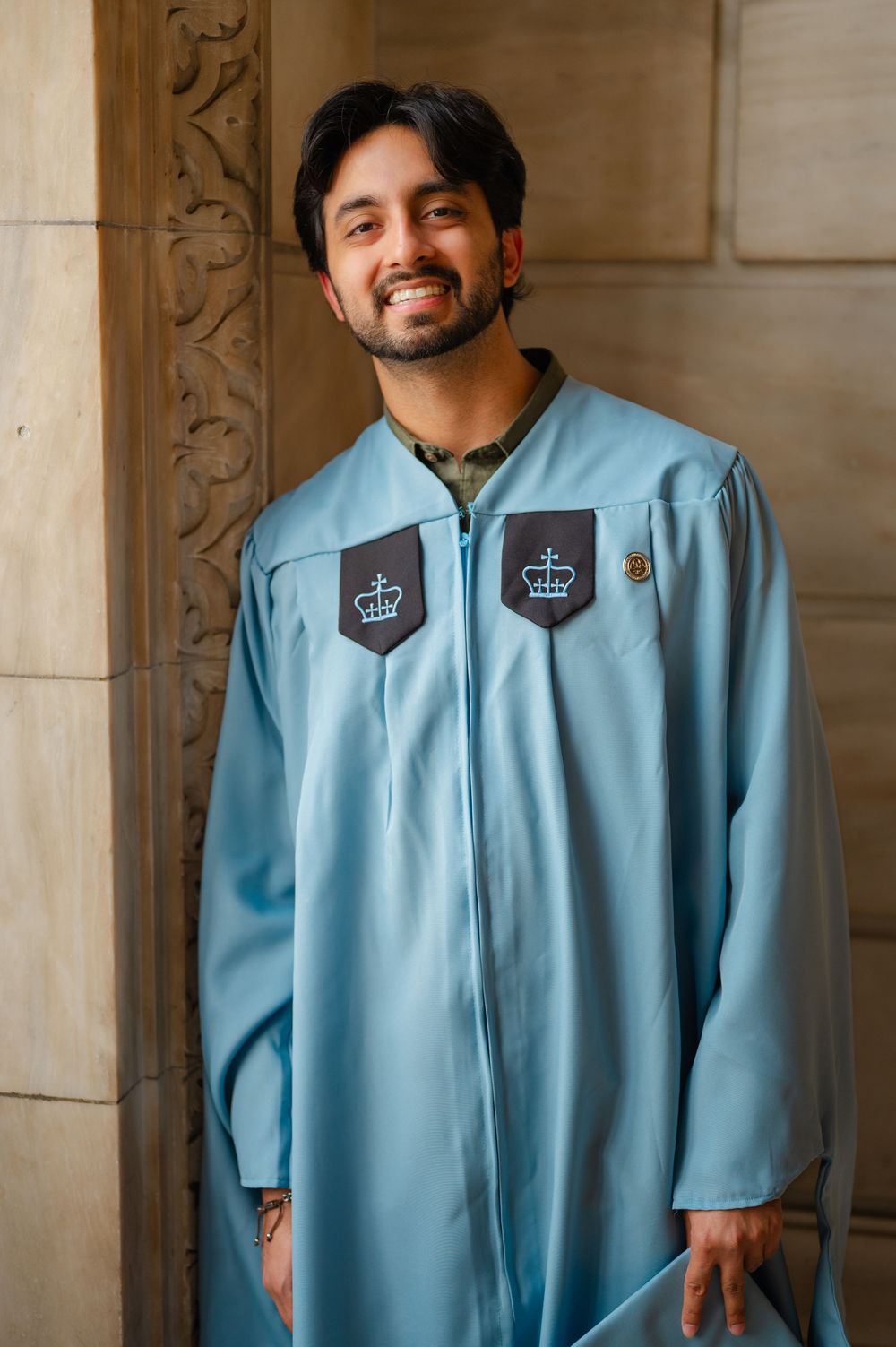 Smiling man in light blue graduation gown with crown emblems posing by ornate stone column.