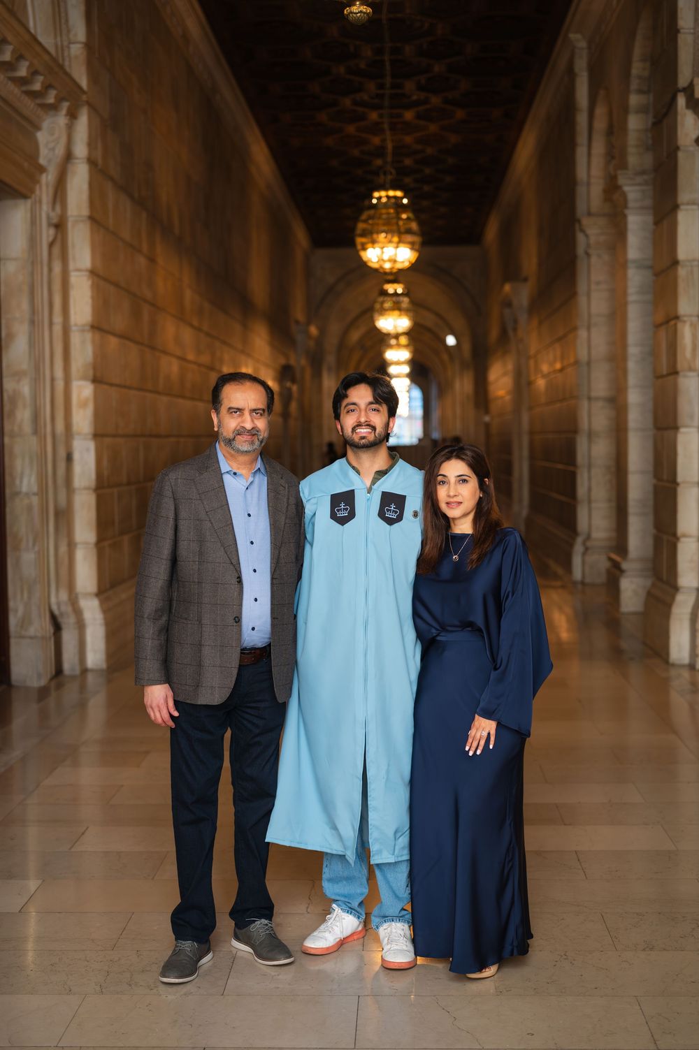 Family of three posing in ornate hallway, young man in light blue graduation gown flanked by parents in formal attire.