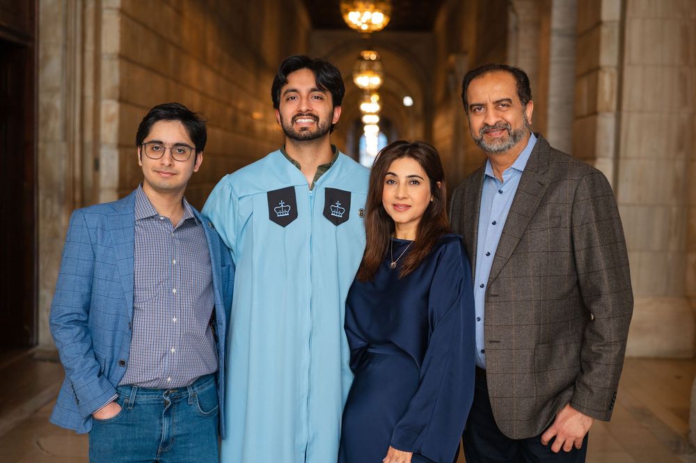 Close-up of smiling family of four celebrating graduation in ornate corridor, graduate in blue robe.