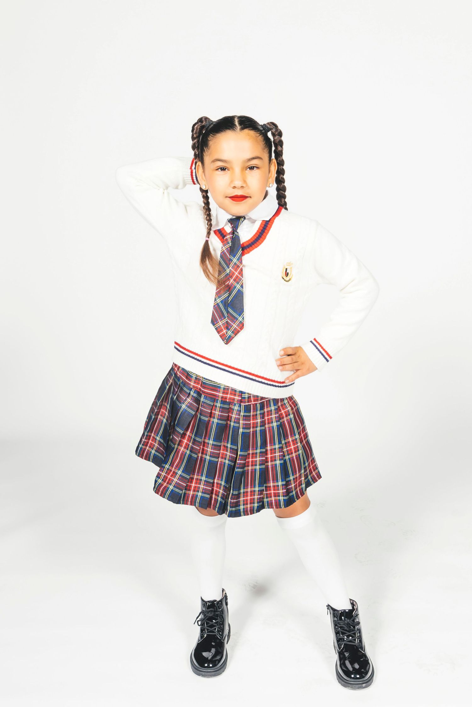 Young girl in school uniform with plaid skirt, white sweater, tie, white socks, and black patent shoes, posing confidently.
