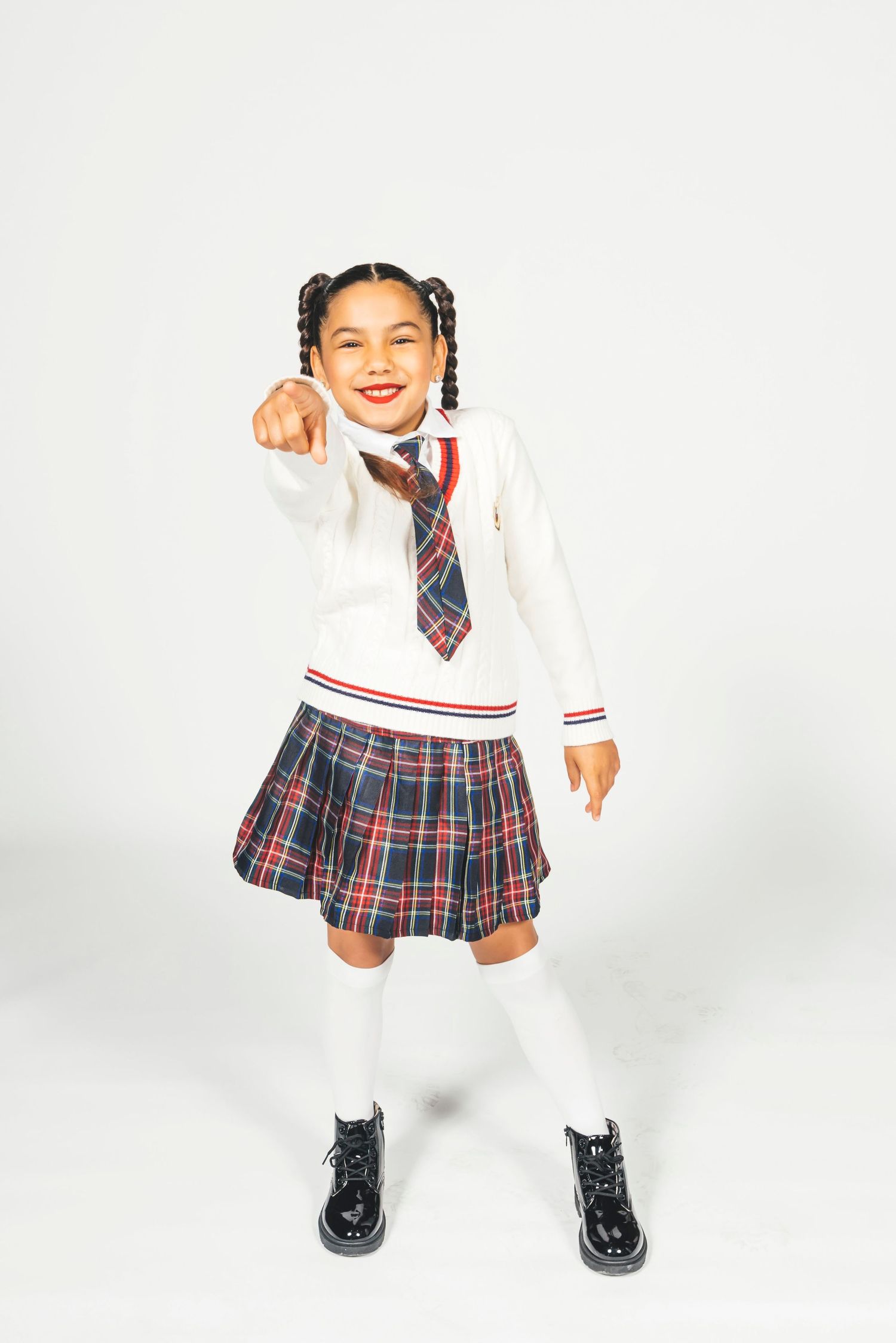 Young girl in plaid school uniform with white sweater, tie, knee socks, and black boots, pointing and smiling.