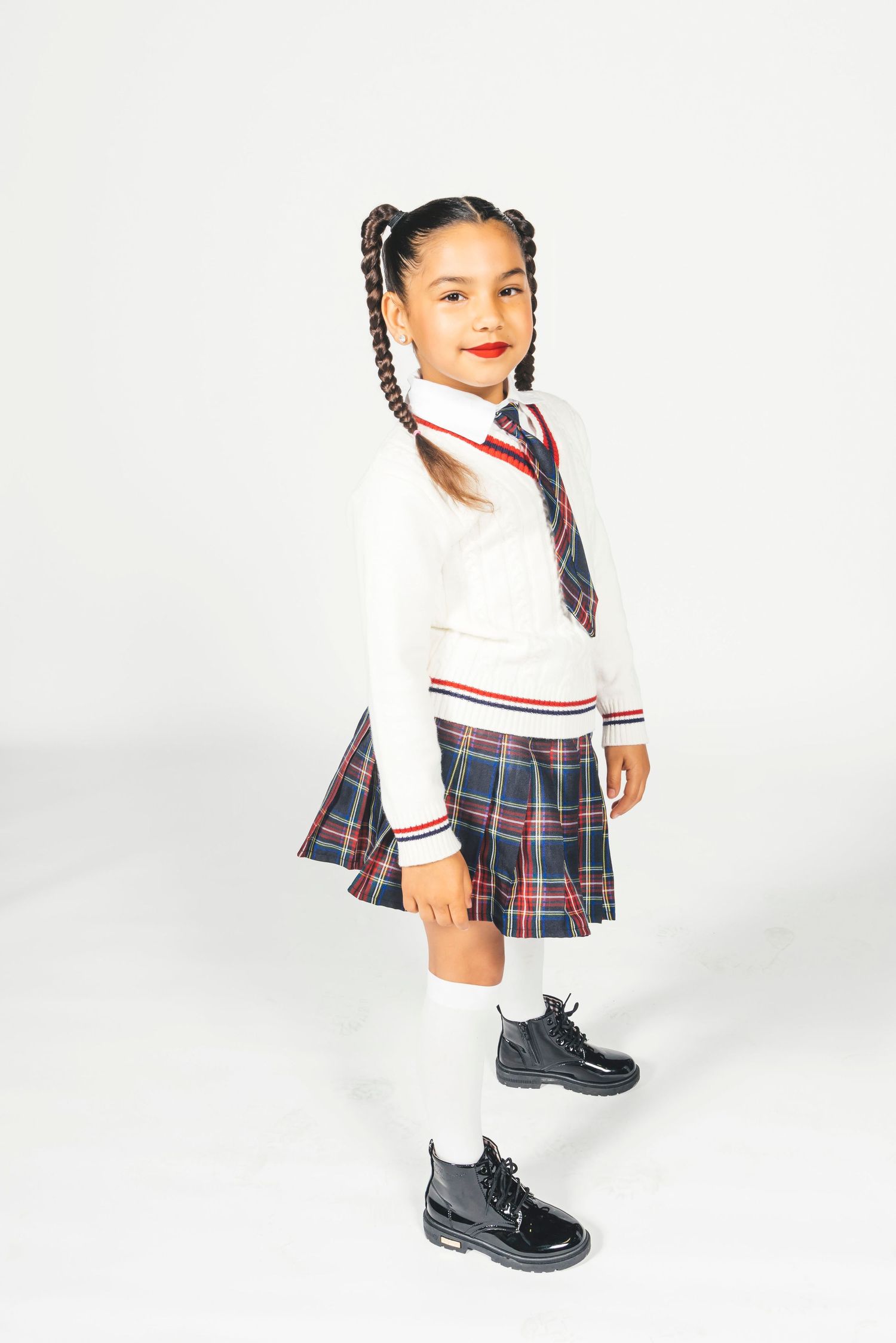 Young girl posing in plaid pleated skirt, white sweater, necktie, knee-high socks, and black patent leather boots.