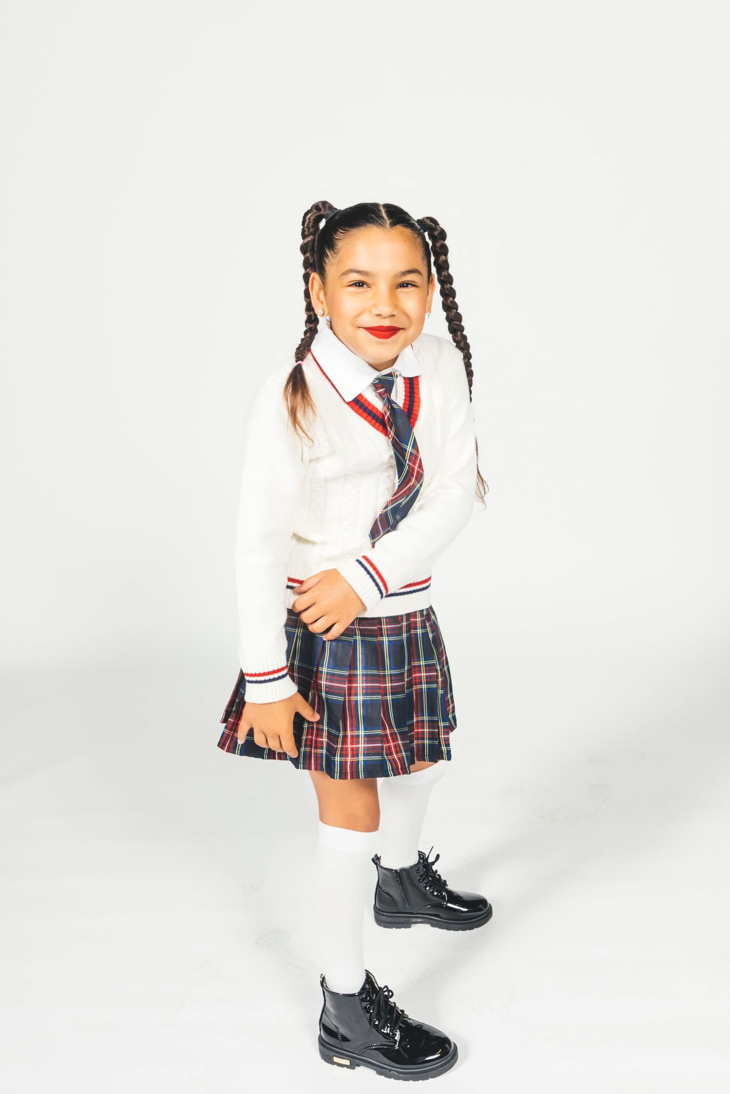 Young girl in plaid school uniform with white sweater, tie, knee socks, and black boots, smiling on white background.
