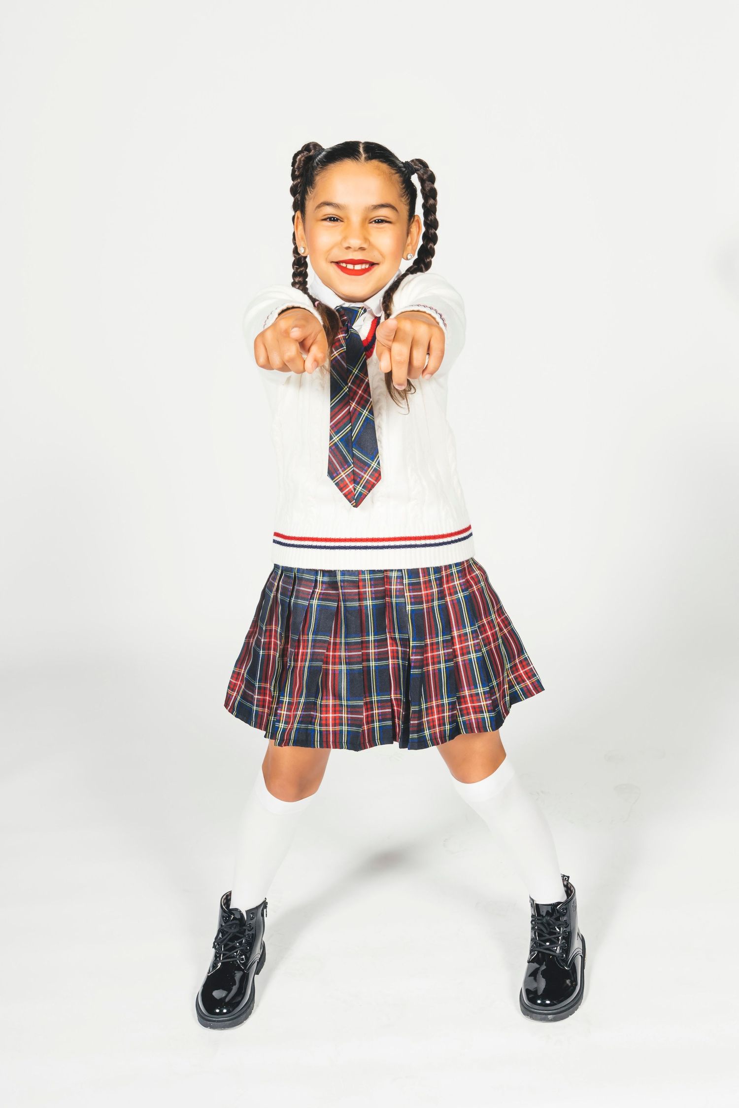 Smiling girl in plaid school uniform skirt, white sweater, tie, knee socks, and black patent boots pointing forward.