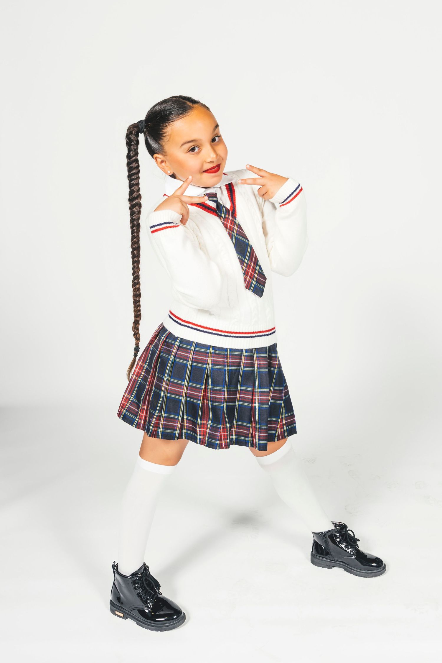 Young girl in school uniform with plaid skirt, white sweater, knee socks, and black shoes, posing playfully.