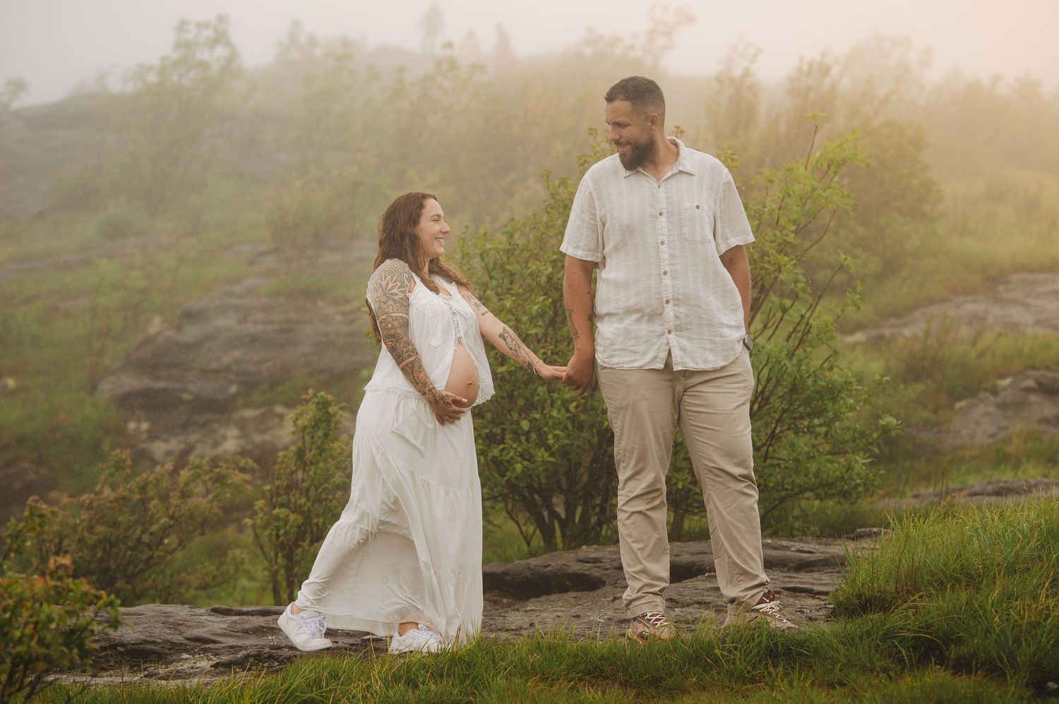 Pregnant couple holding hands on misty rocky hillside, woman in white maternity gown, man in white shirt and khaki pants.