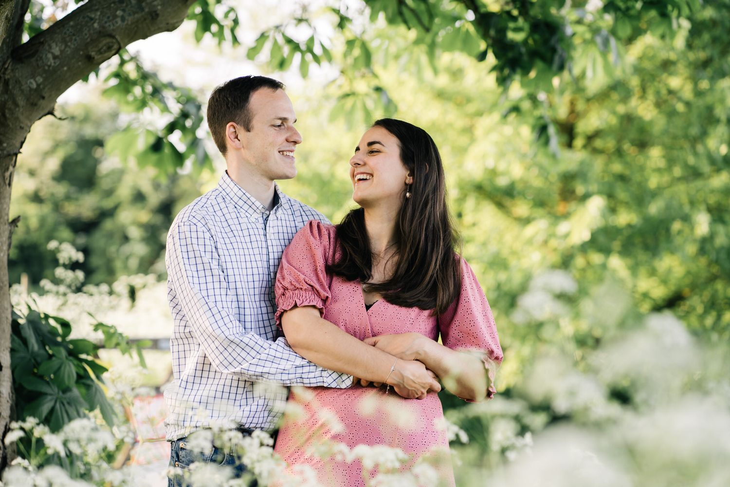 Happy couple embracing and smiling outdoors among lush green trees and white flowering bushes on a sunny day.