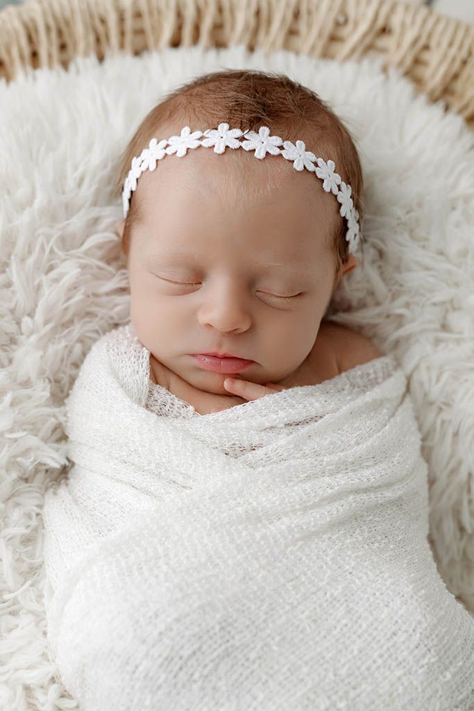 Sleeping newborn baby girl swaddled in white wrap with floral headband resting in a basket.