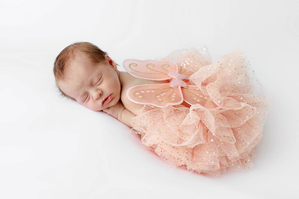Sleeping newborn baby dressed in a pink tutu and fairy wings on a white background.