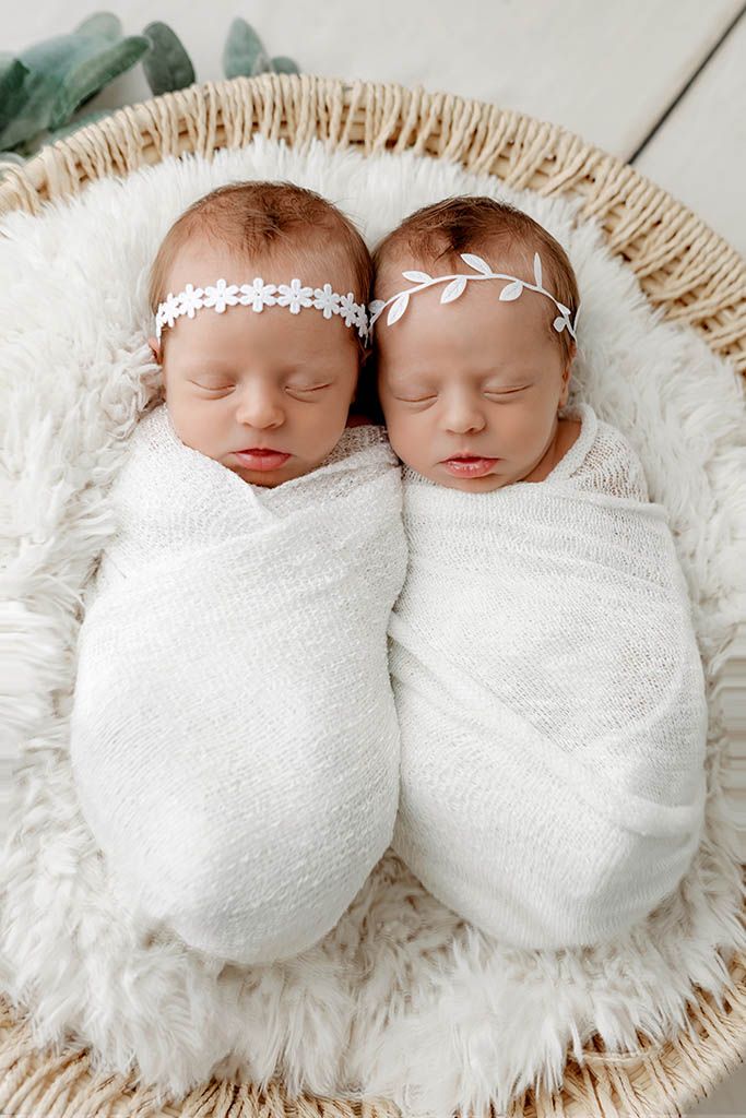 Two sleeping newborn twin girls swaddled in white wraps, wearing floral headbands by Murrieta newborn twins photographer.