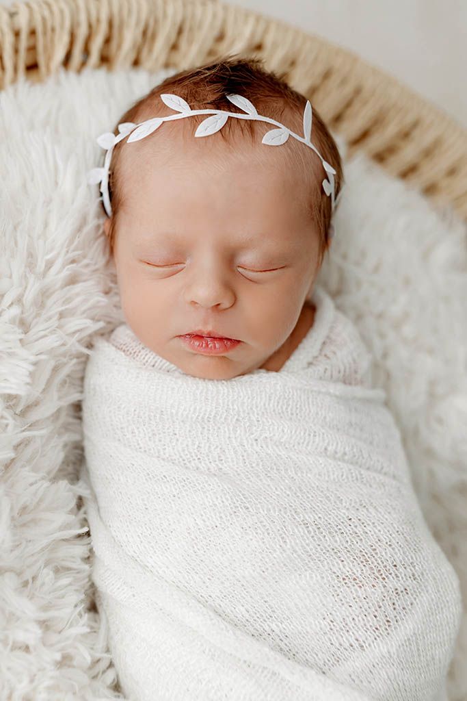 Sleeping newborn baby swaddled in white knit wrap wearing a delicate leaf headband, resting in a wicker basket.