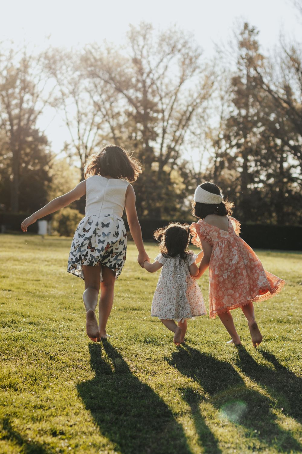 Three young girls in summer dresses holding hands while running through a sunny green park at golden hour.