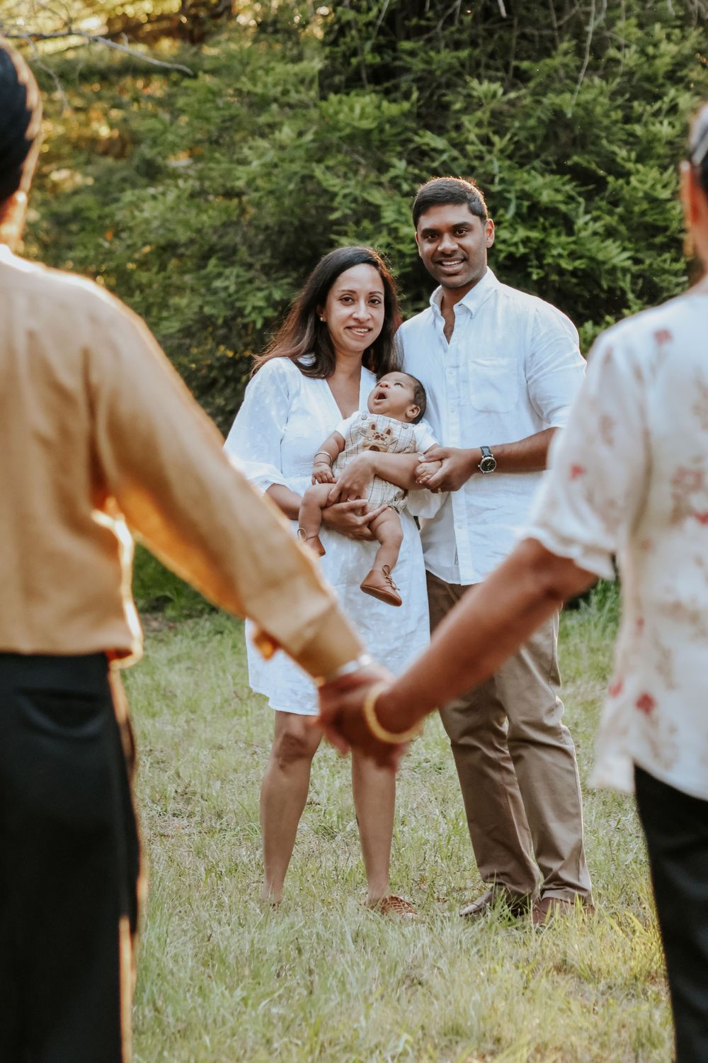 A smiling couple holds their baby outdoors on grass with green trees behind them as two people stand in the foreground.