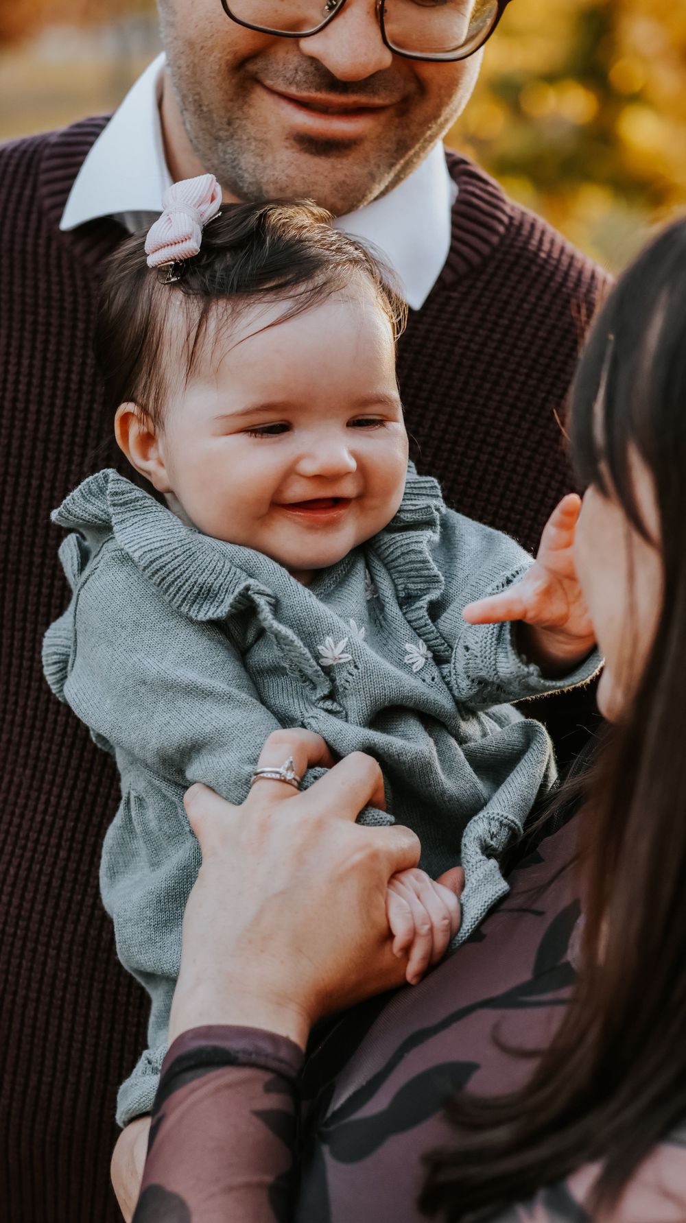 A smiling baby girl in a green ruffled sweater with a pink bow is held by her parents outdoors in autumn.