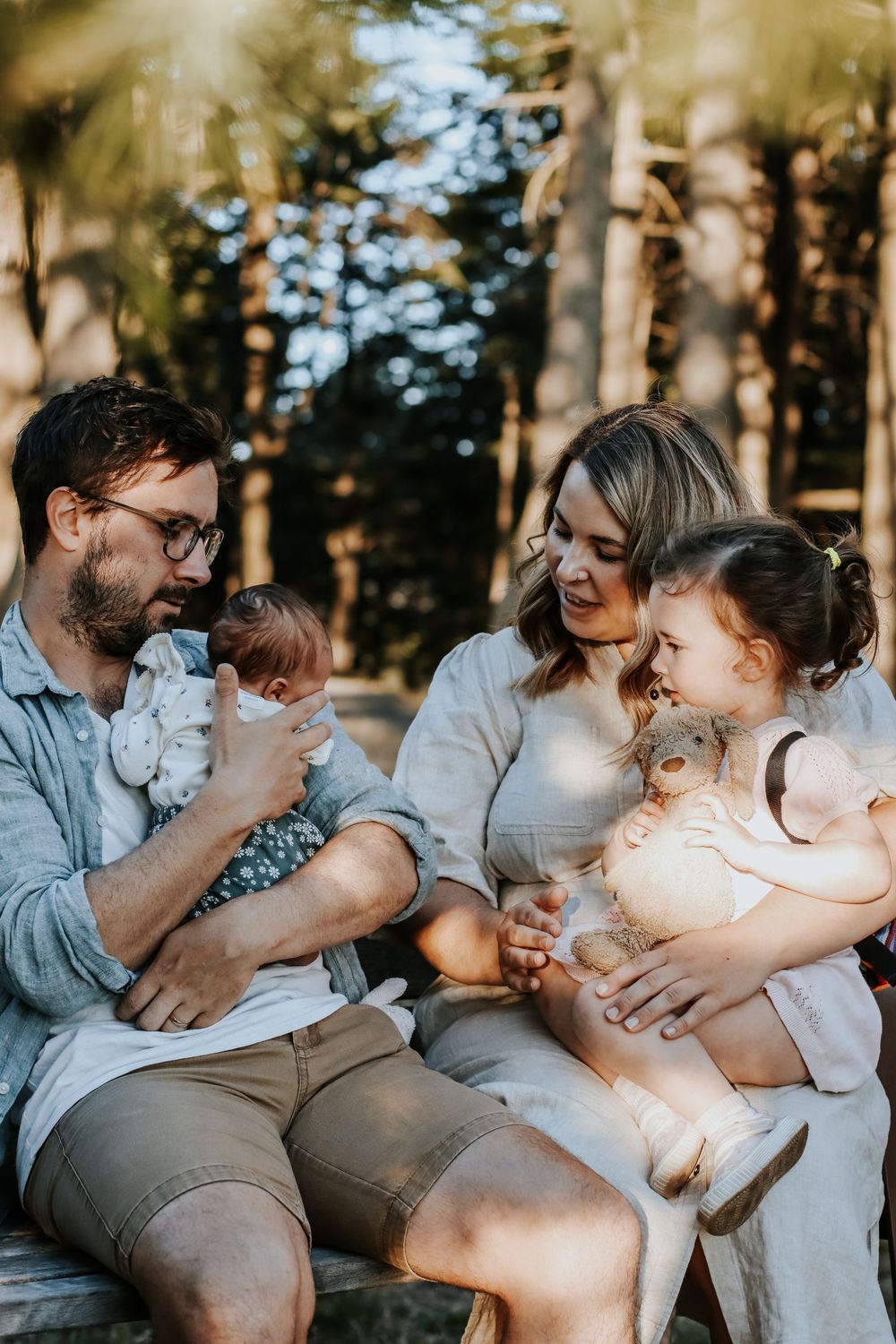 Family of four sitting outdoors in sunlit forest, parents holding baby and toddler with teddy bear.