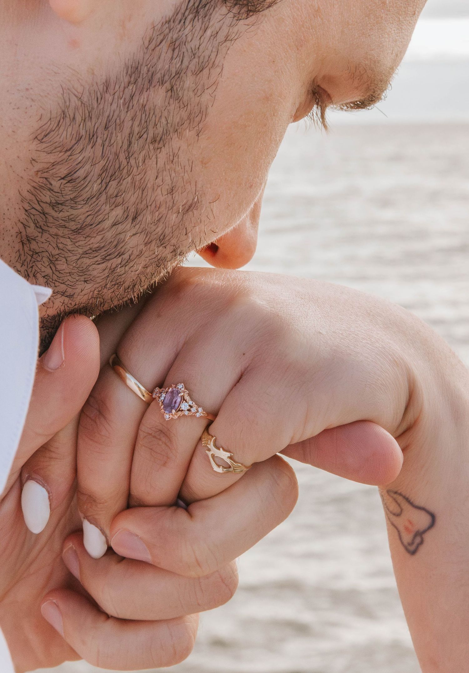 Man kissing woman's hand adorned with gold engagement ring featuring purple gemstone, near beach.