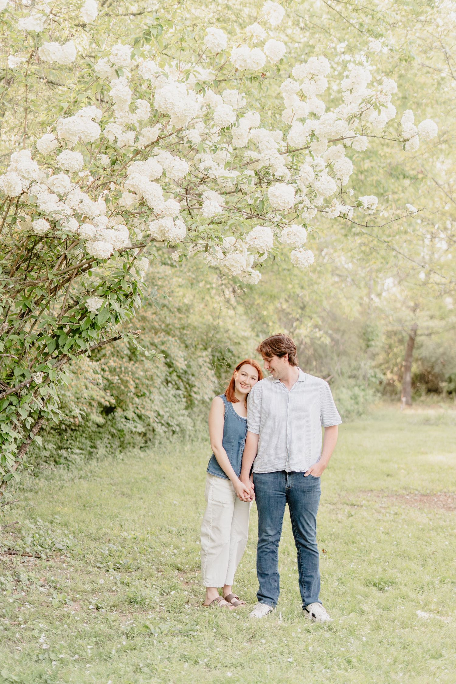 Couple holding hands under blooming white flowers in a lush green park, wearing casual spring outfits.