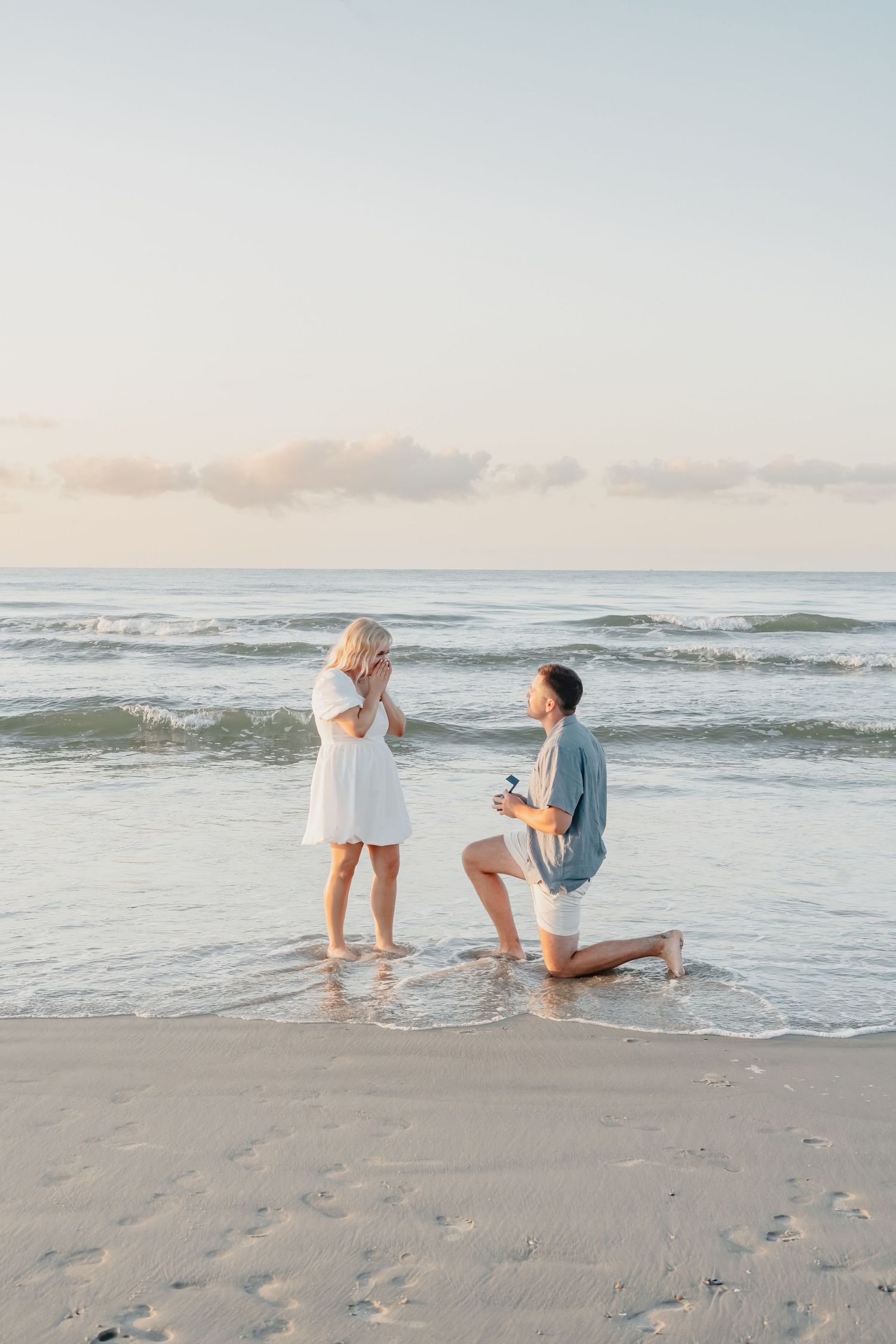 Man proposes on bended knee to woman on a sandy beach at sunset, holding a ring box as waves roll in.