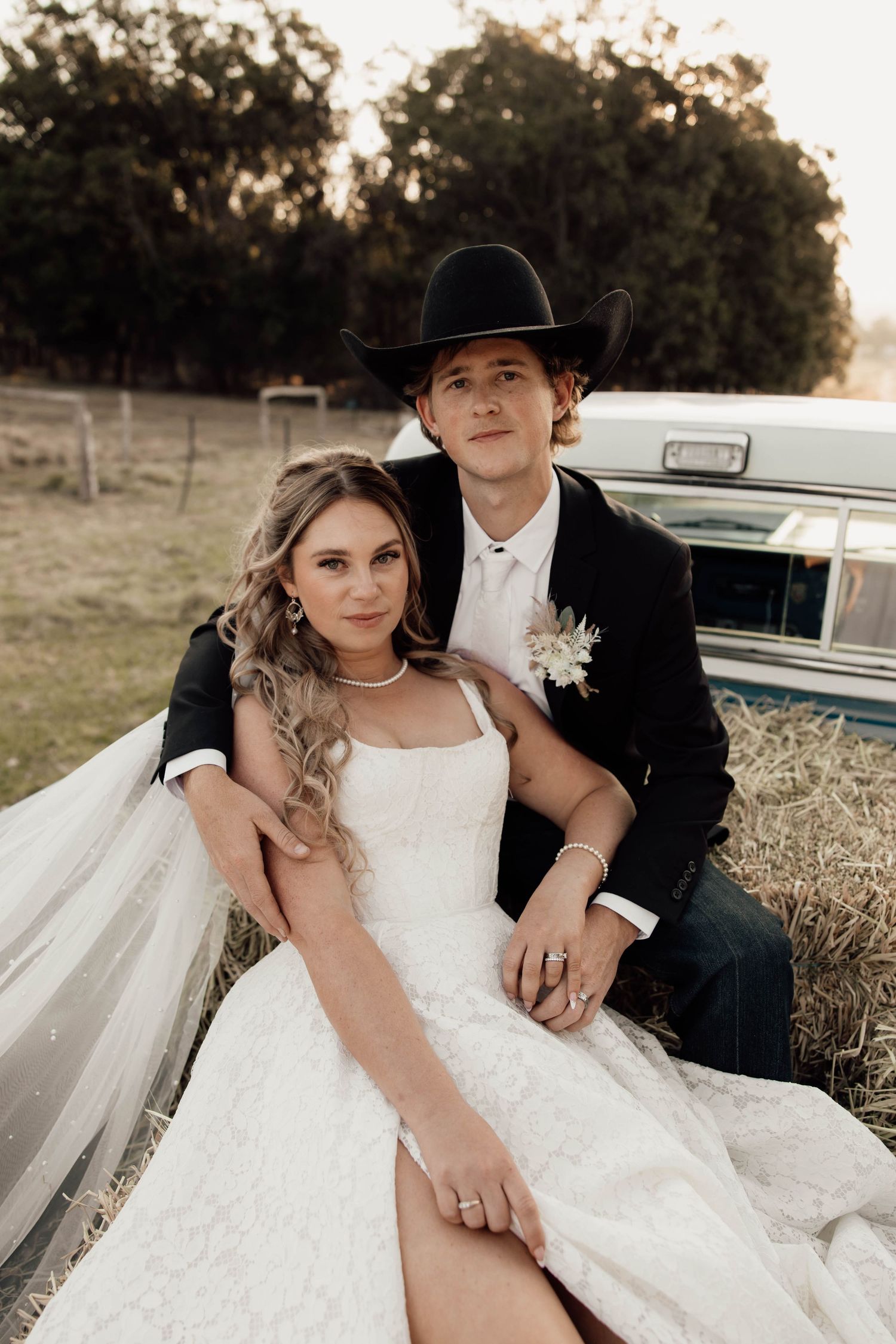 Bride and groom sitting in vintage truck bed at rustic farm wedding, groom wearing black cowboy hat and boutonniere.