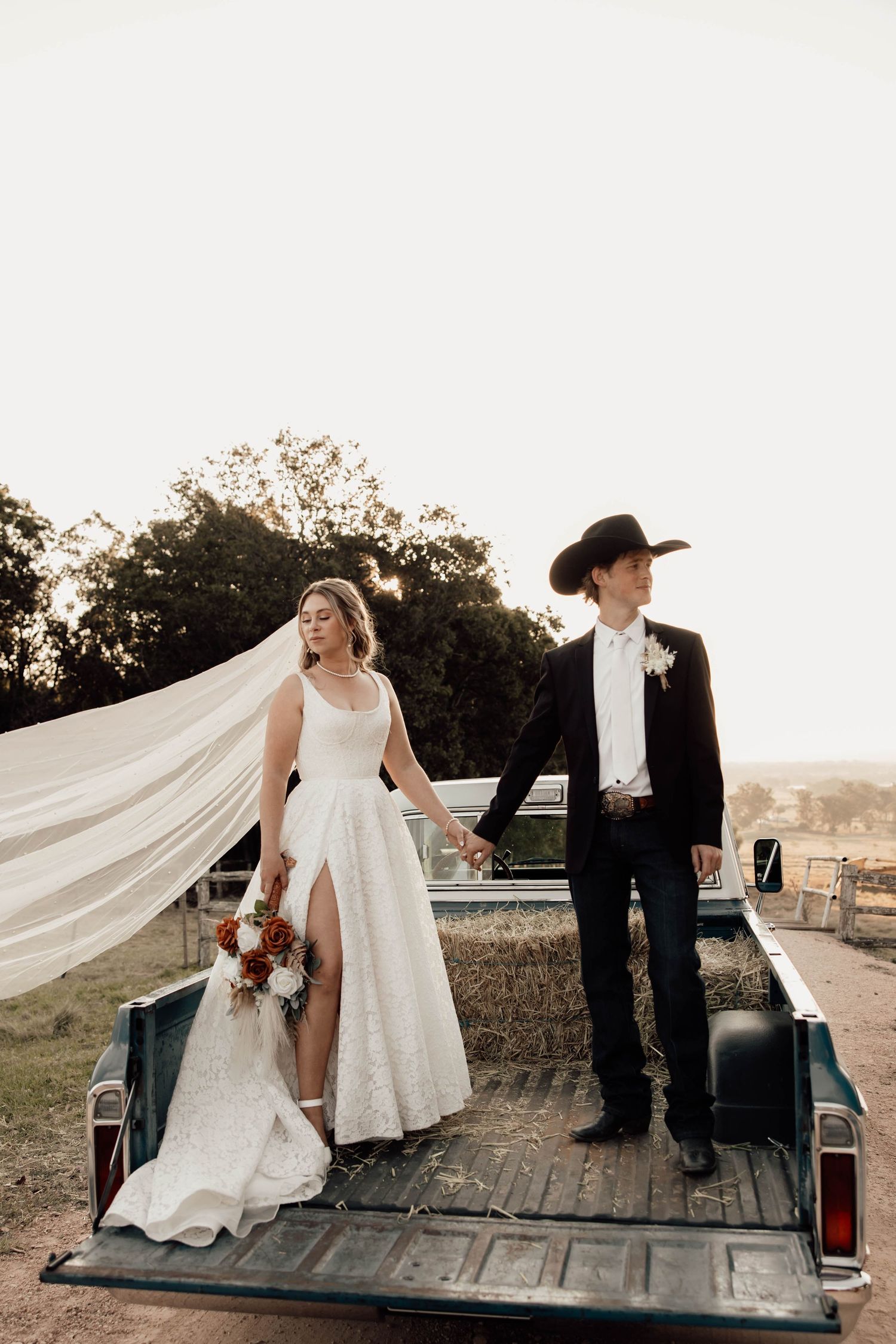 Bride and groom standing in vintage truck bed, bride holding bouquet with veil flowing, groom in black cowboy hat.