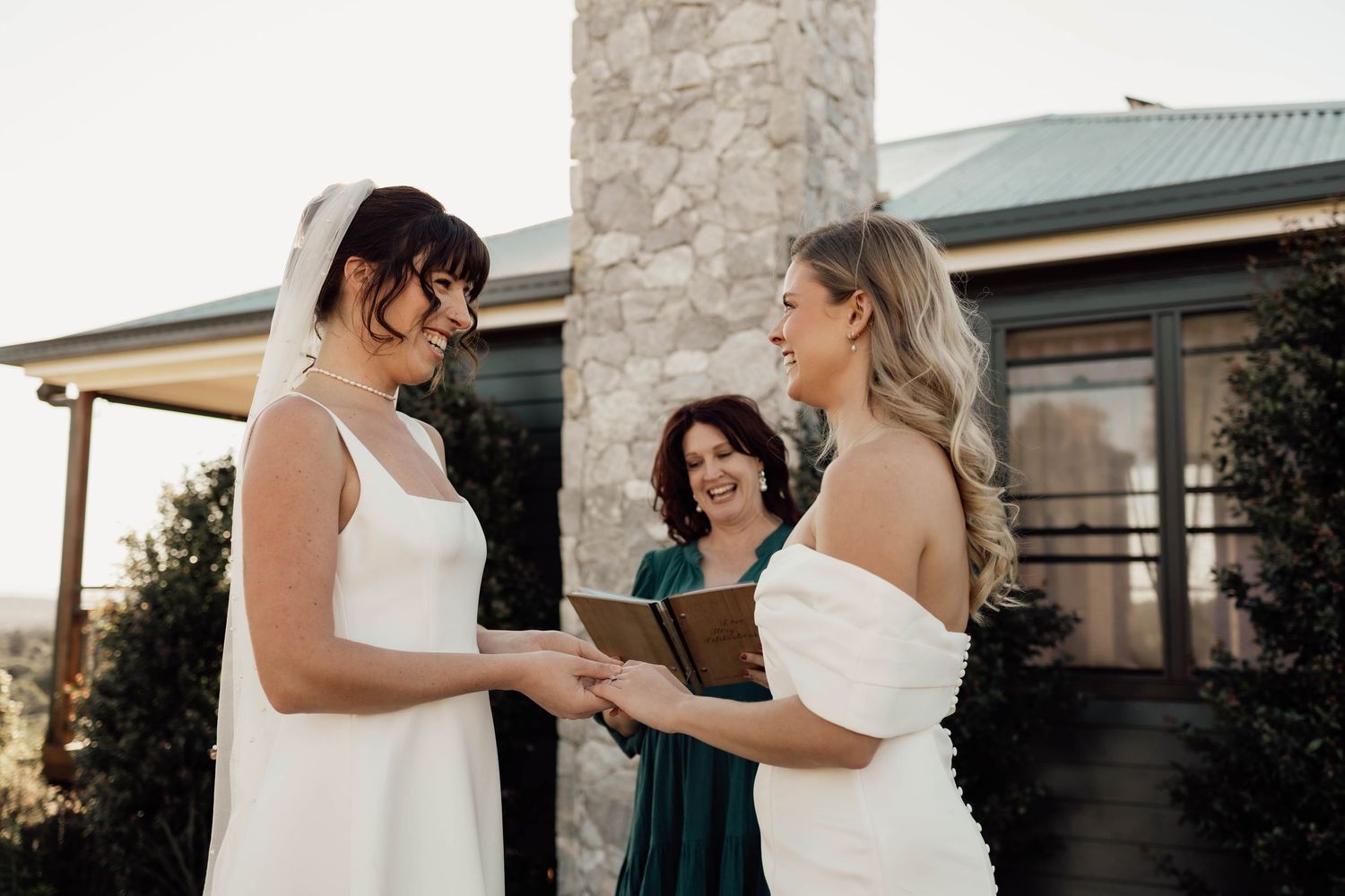 Two brides in white gowns exchange vows outdoors with a female officiant holding a book at a wedding ceremony.