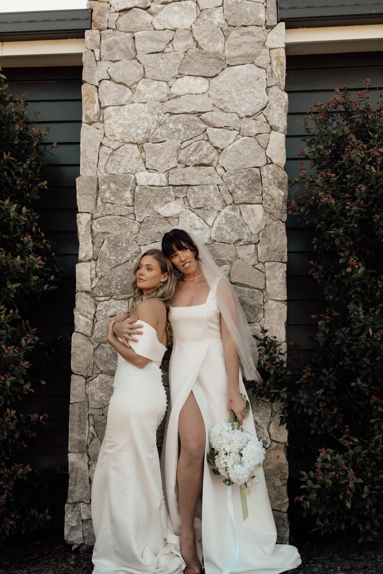 Two brides in elegant white gowns pose together before a stone wall, one holding a white floral bouquet.