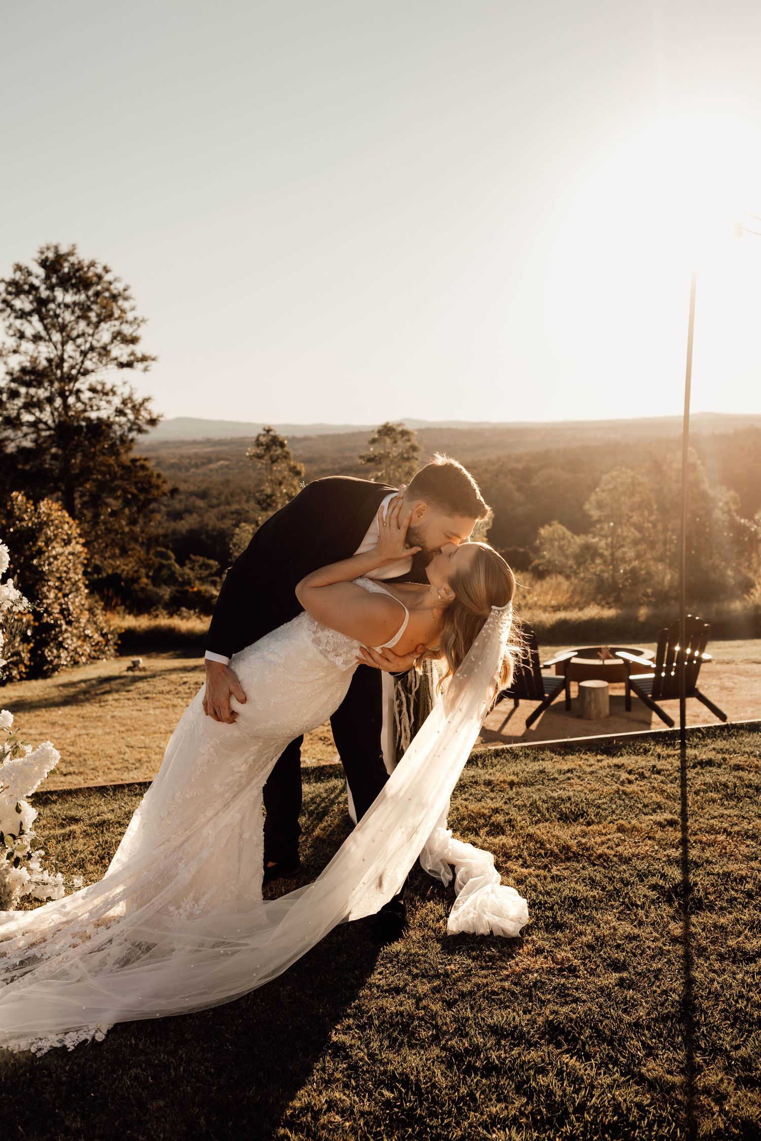 Bride and groom share a romantic dip kiss at golden hour with scenic rolling hills backdrop at outdoor wedding venue.