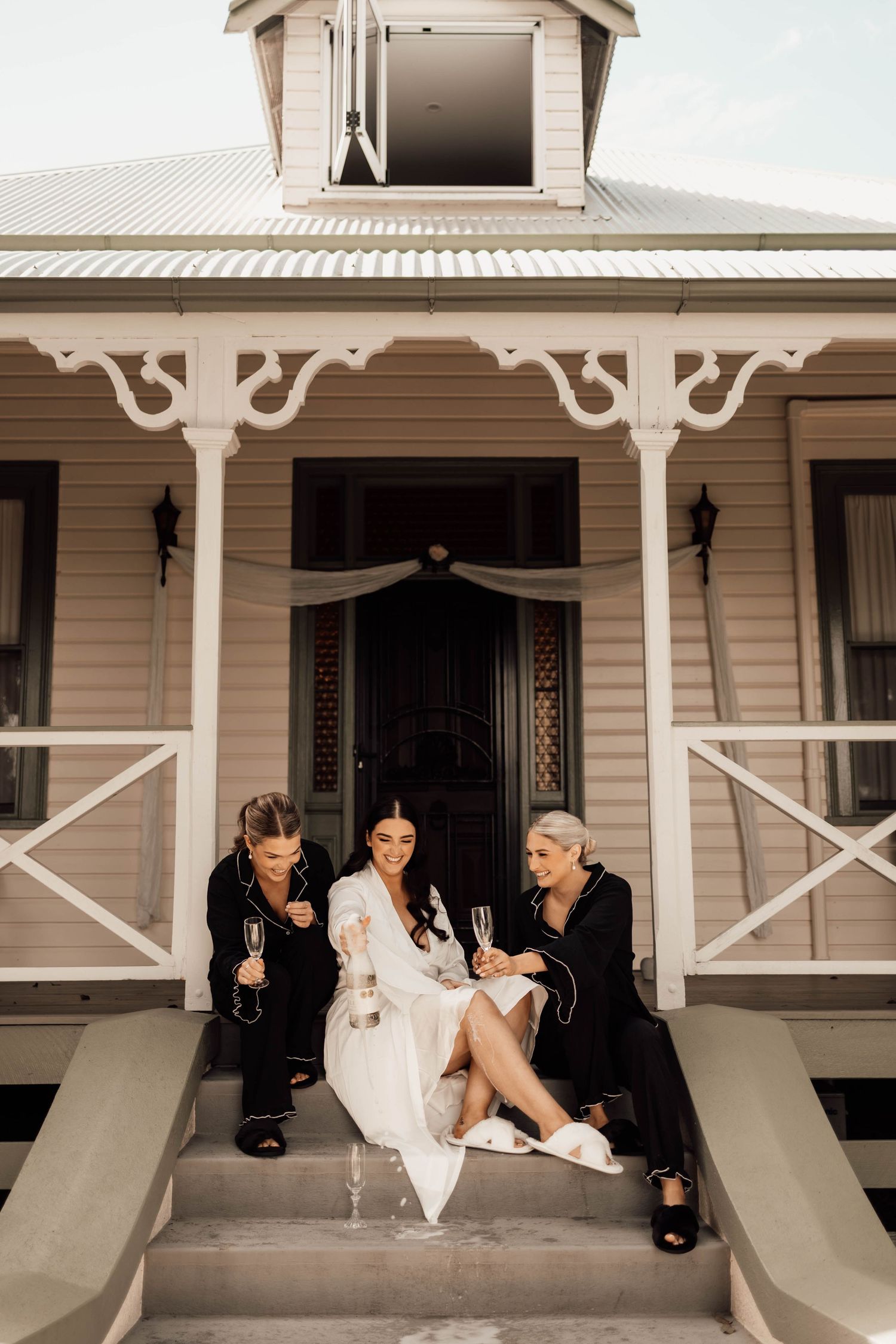 Bride in white robe celebrates with two bridesmaids in black on a charming vintage house porch, holding champagne glasses.