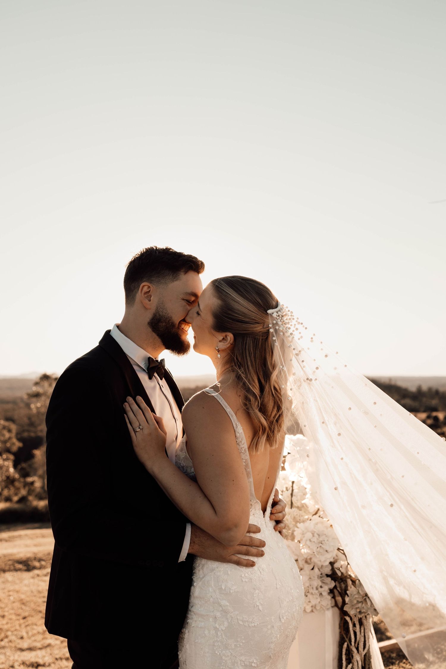 A bride and groom share a romantic moment outdoors at golden hour, with her veil flowing in the breeze.