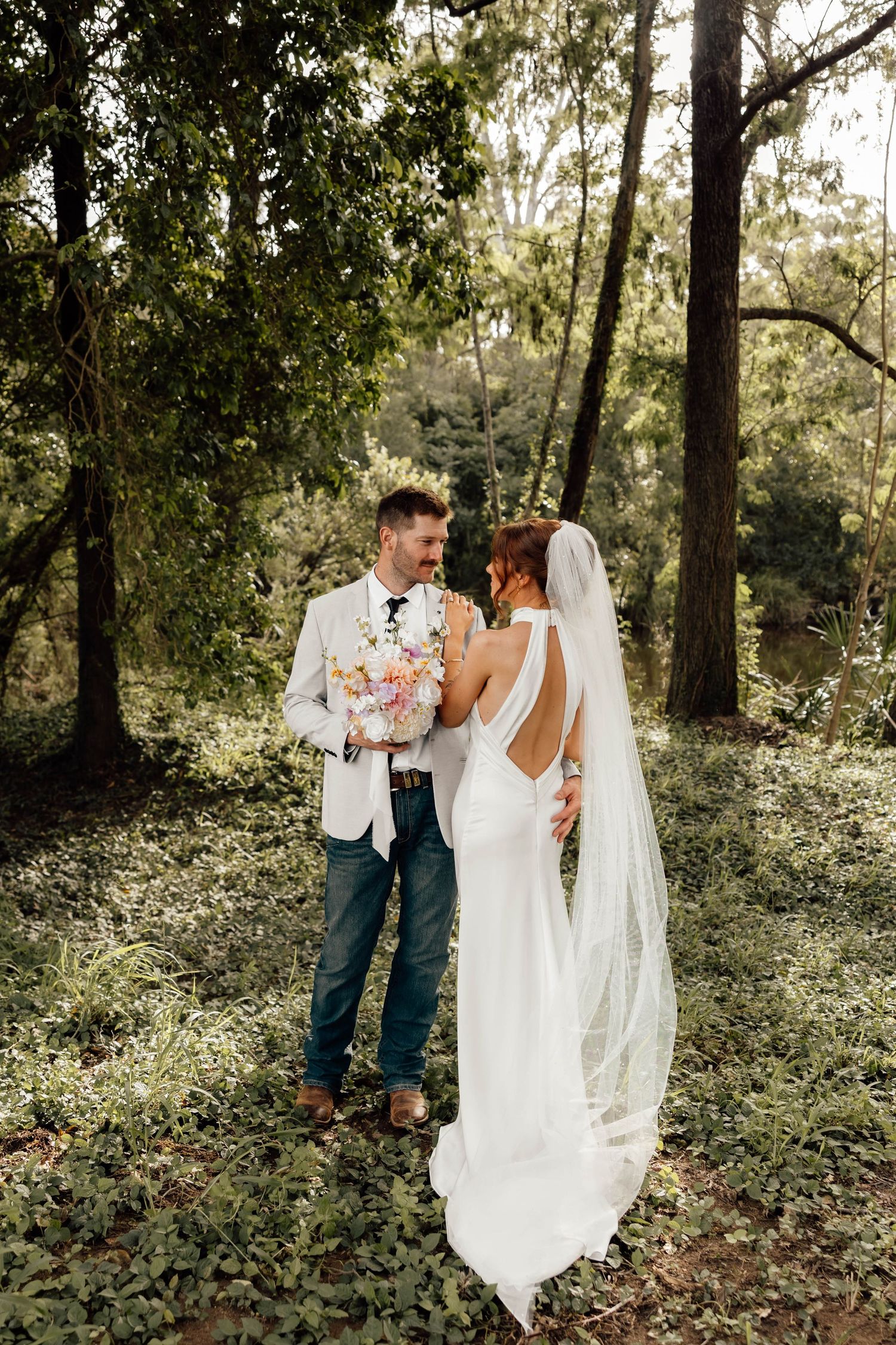 A bride and groom share a romantic moment in a lush forest setting, with the bride in a backless white gown and veil.