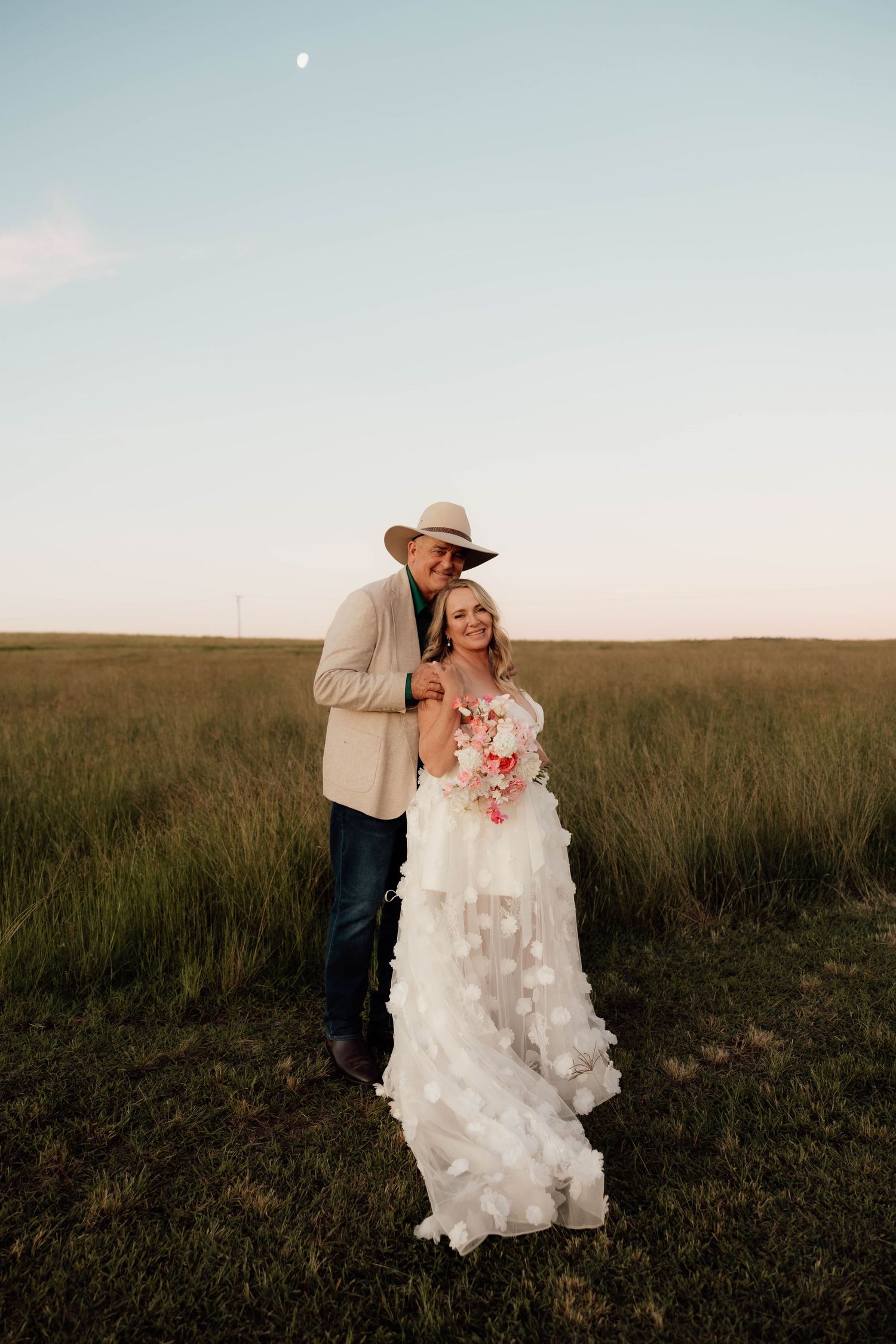 Bride in white floral gown holding bouquet with groom in cowboy hat embracing in green field at sunset.