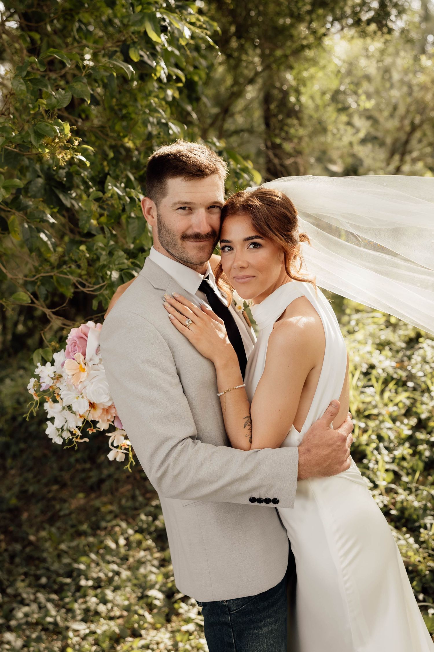 A bride and groom embrace outdoors, she wears a flowing veil and white gown, he holds a floral bouquet in a beige suit.