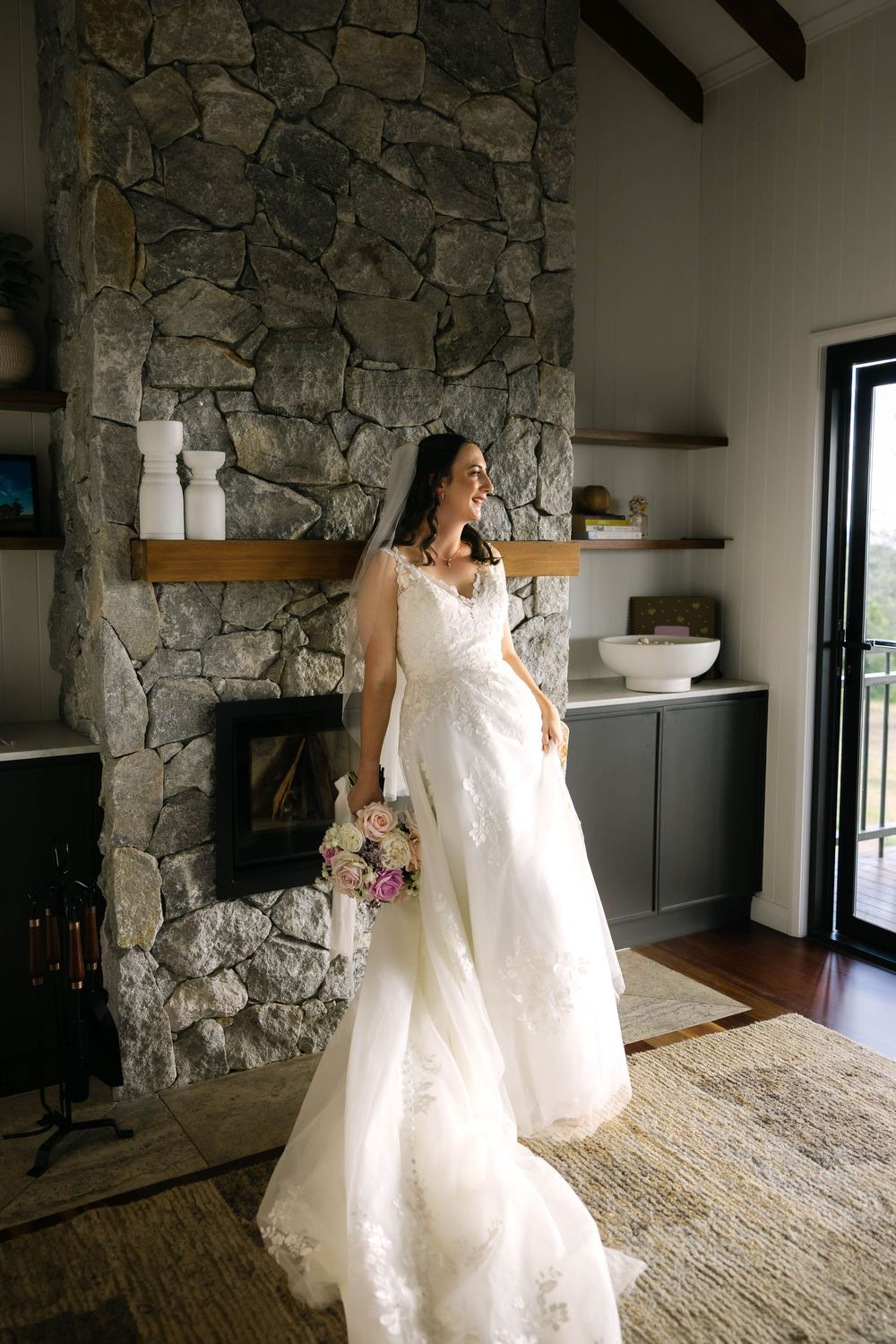 Bride in white lace gown holding bouquet stands before stone fireplace in elegant rustic interior.