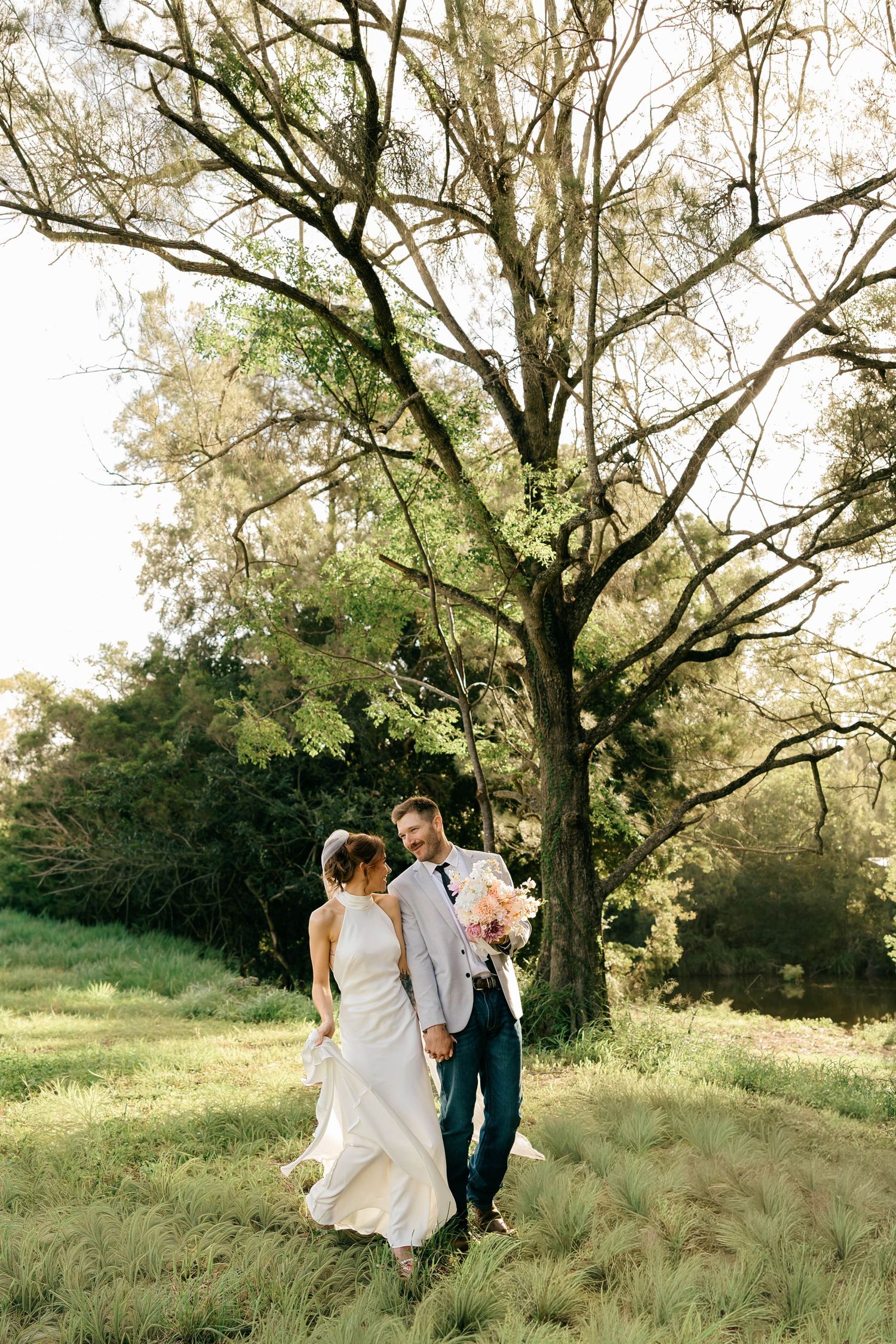 A bride and groom walk hand in hand through a lush green field beneath a large tree on their wedding day.