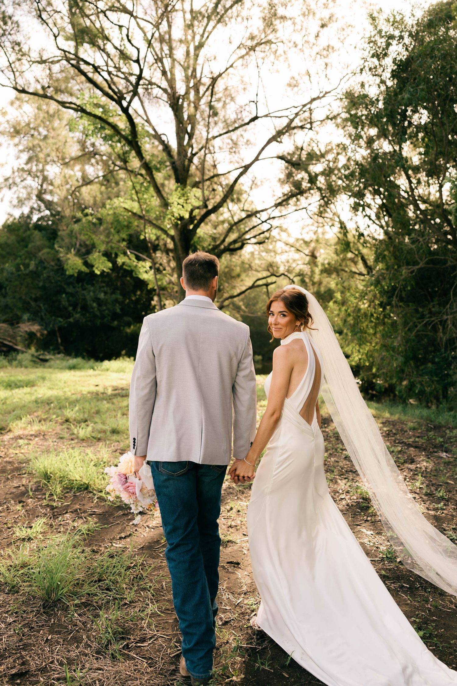Bride and groom walking hand in hand through a lush green outdoor setting, bride wearing a white gown with long veil.