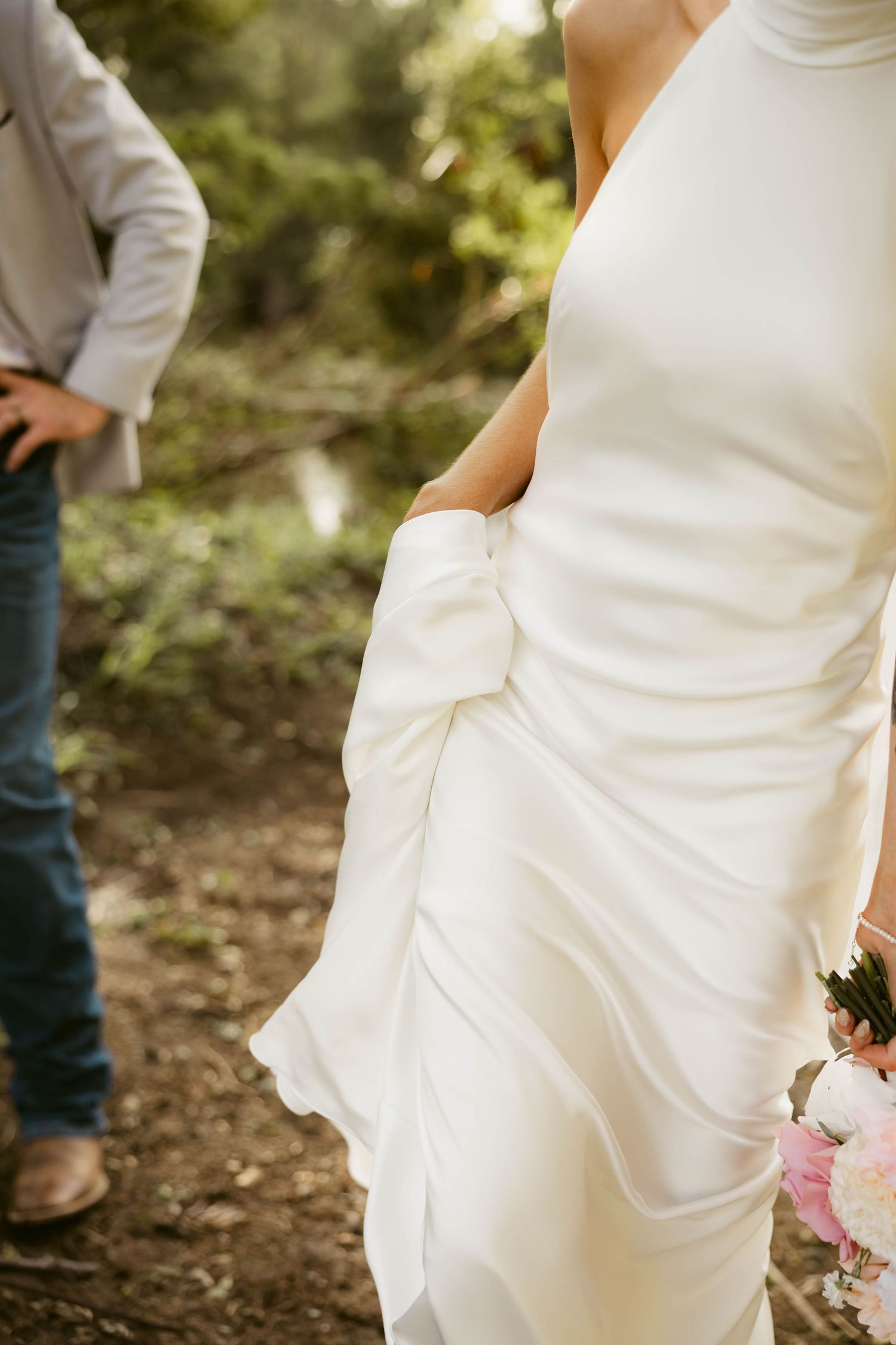 Bride in elegant white wedding gown holding bouquet of pink flowers outdoors, with groom in jeans visible in background.