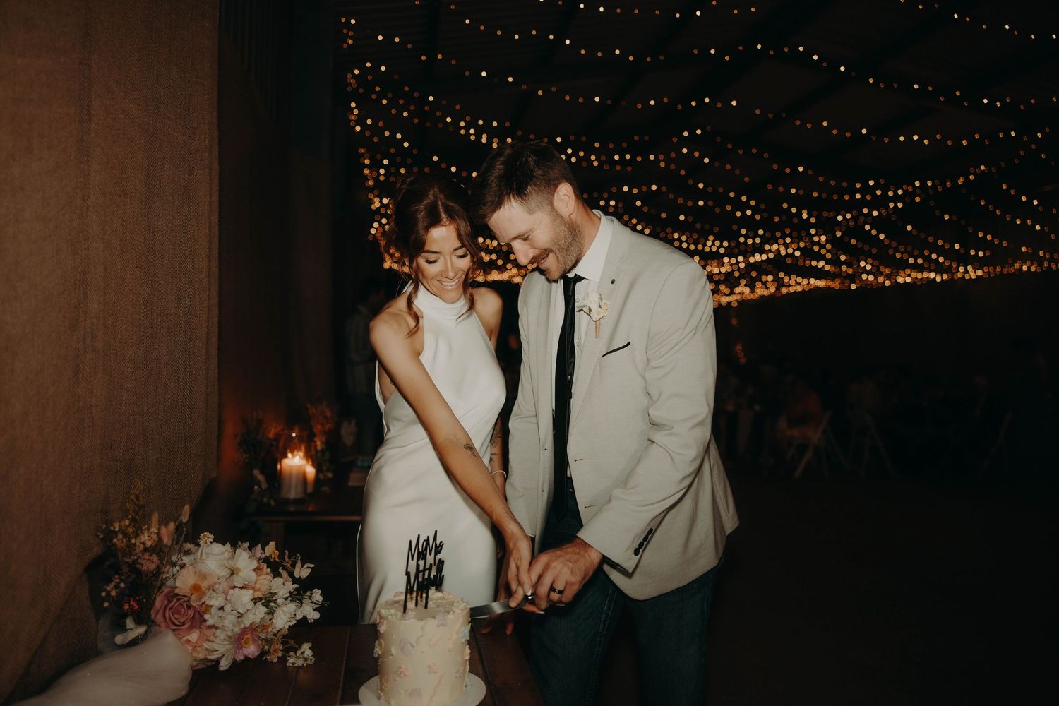 Bride and groom cutting white wedding cake under fairy lights at romantic evening reception.