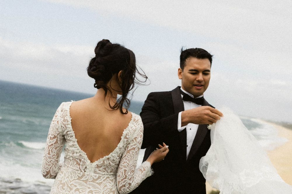 Bride in lace backless gown and groom in tuxedo share romantic beach wedding moment with ocean waves in background.