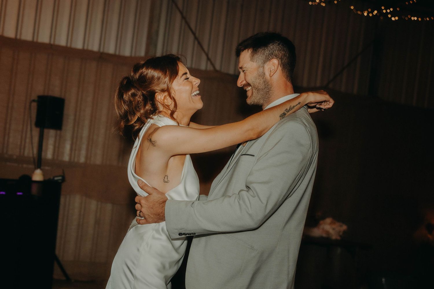 Bride and groom share a joyful first dance, smiling at each other in a warmly lit barn wedding reception.