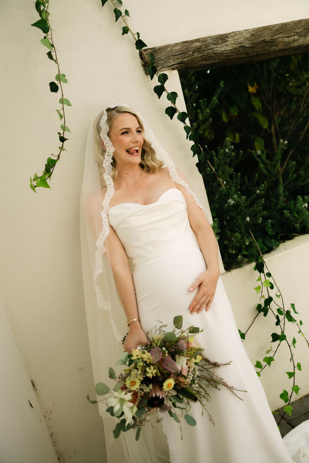 Laughing bride in white strapless gown and lace veil holds wildflower bouquet beside ivy-covered wall.