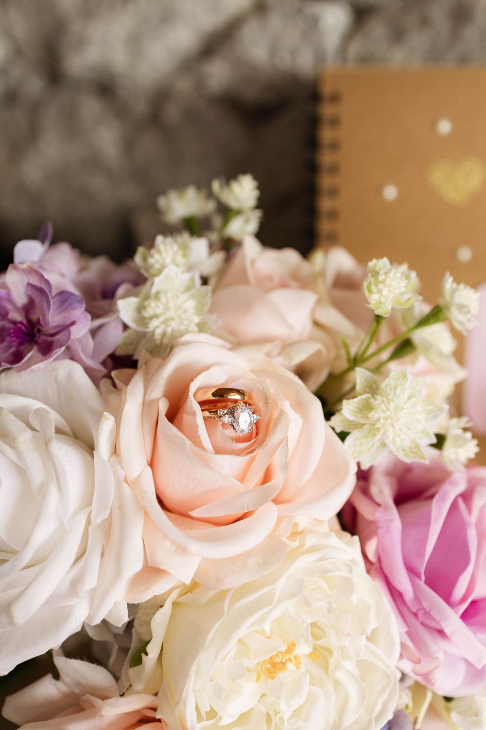 Gold engagement ring resting in a blush pink rose surrounded by a romantic bouquet of white, purple, and pink flowers.