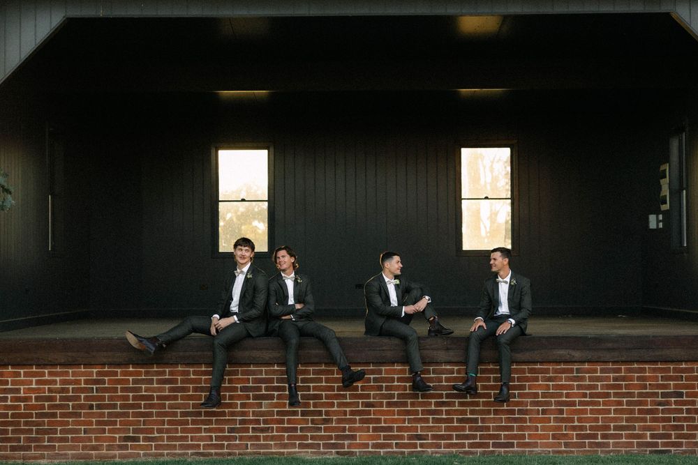 Four groomsmen in dark suits sitting on a brick wall outside a modern black building.