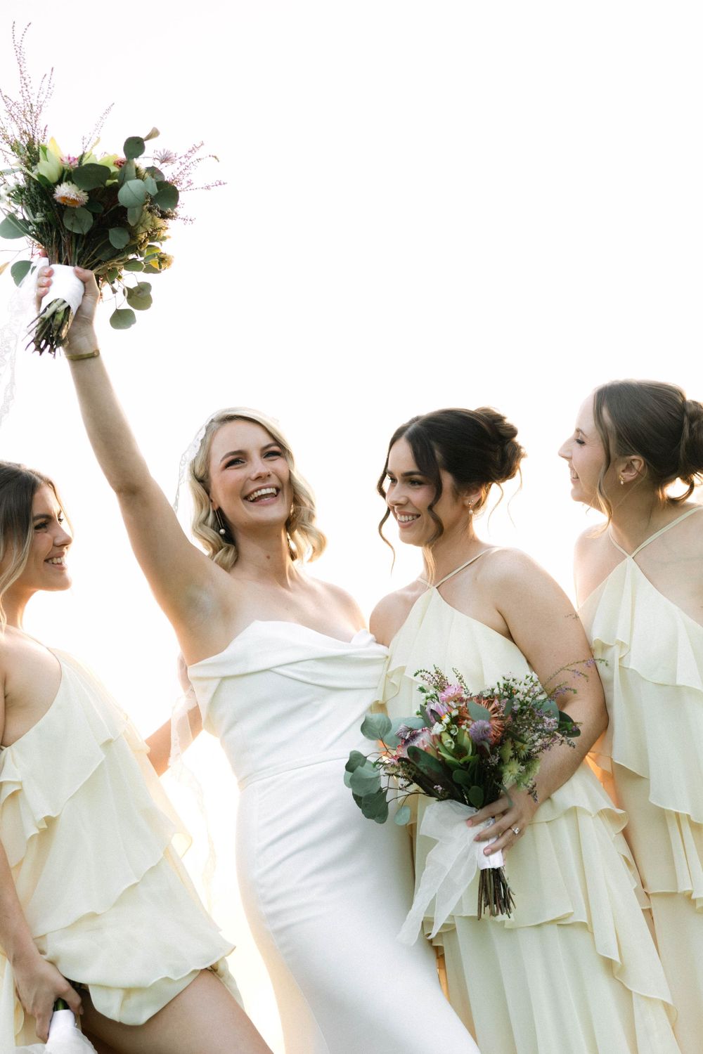 Bride in white gown raises bouquet joyfully with three bridesmaids in cream ruffled dresses holding floral arrangements.