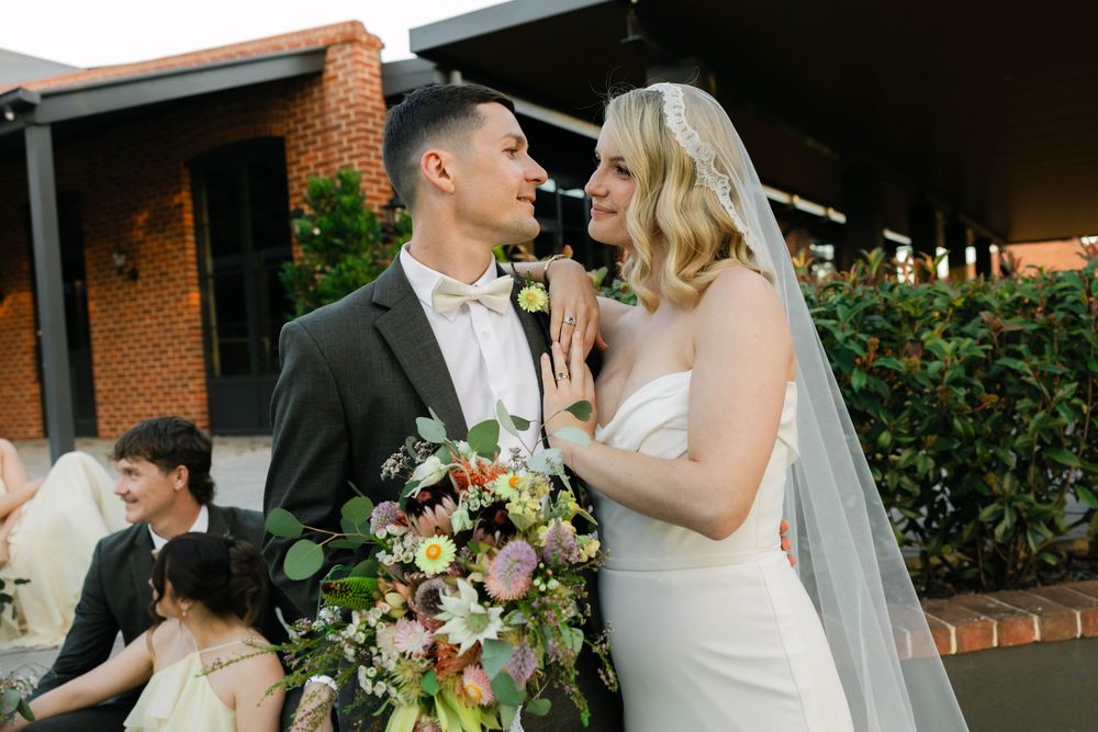 Smiling bride and groom share a romantic moment outdoors, bride holding a lush floral bouquet.